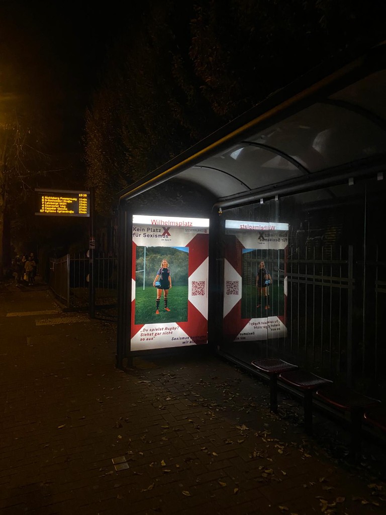 Beleuchtetes City-Light an der Bushaltestelle Wilhelmsplatz mit Rugby-Motiv der Kampagne „Kein Platz für Sexismus im Sport“, daneben die digitale Abfahrtstafel