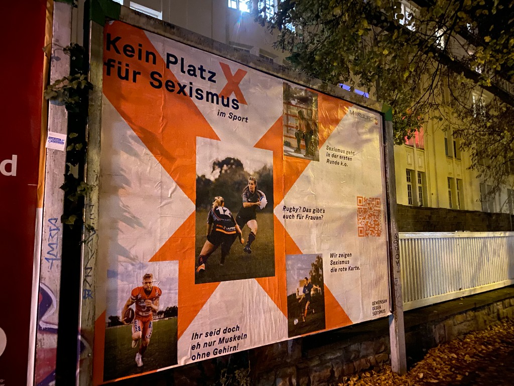 Beleuchtete Großfläche nachts: Kampagne „Kein Platz für Sexismus im Sport“ mit Rugby, Football und QR-Code