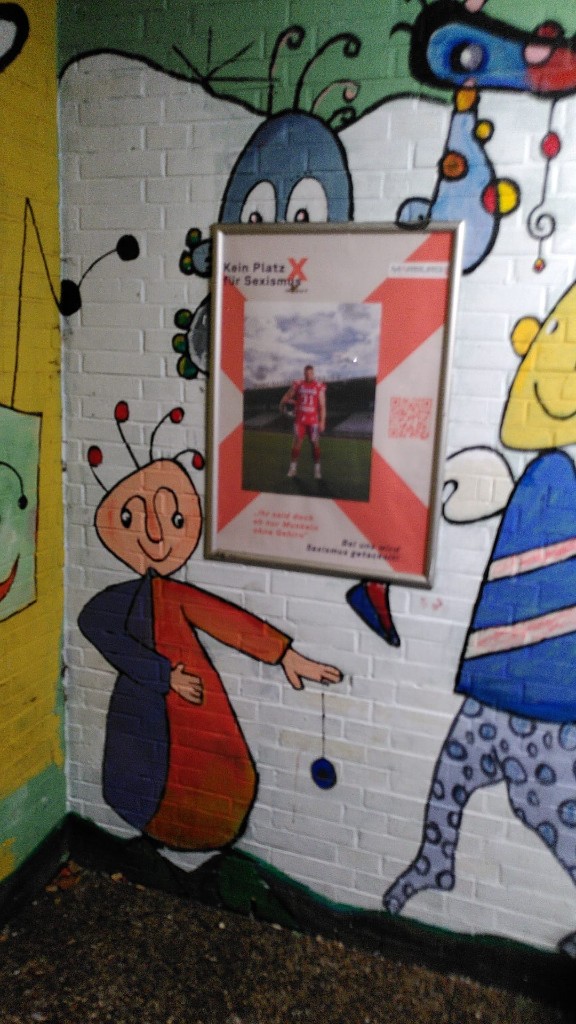 Gerahmtes Plakat „Kein Platz für Sexismus“ mit Football-Spieler der Marburg Mercenaries an einer bemalten Ziegelwand in Marburg