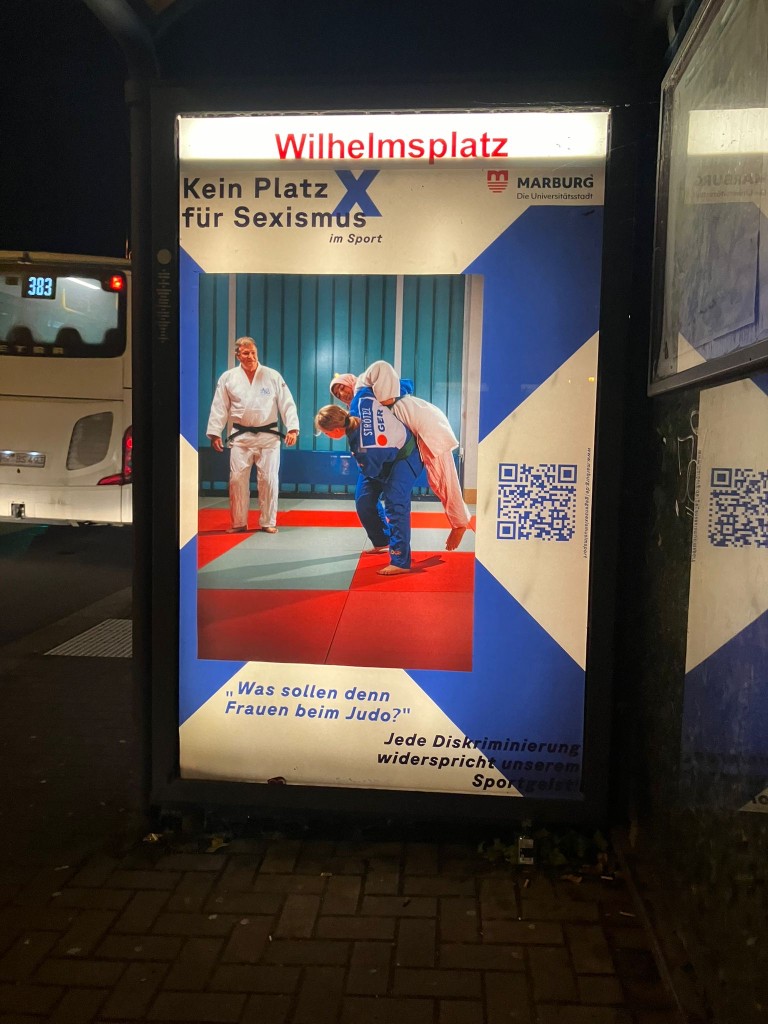 Beleuchtetes Kampagnenplakat mit Judo-Motiv an einer Bushaltestelle am Wilhelmsplatz in Marburg bei Nacht